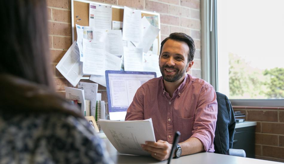 The image shows a male advisor sitting at his desk, smiling and holding a document, engaged in a conversation with a student whose back is facing the camera. The advisor is wearing a pink button-up shirt and is seated in an office with a brick wall, a window, and a bulletin board filled with papers. The setting appears casual and welcoming, indicating a supportive and productive advising session. The sunlight coming through the window suggests a bright and positive atmosphere.