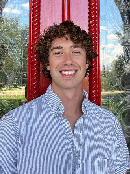 August Railey wears a blue and white striped button up shirt and stands in front of the red doors.