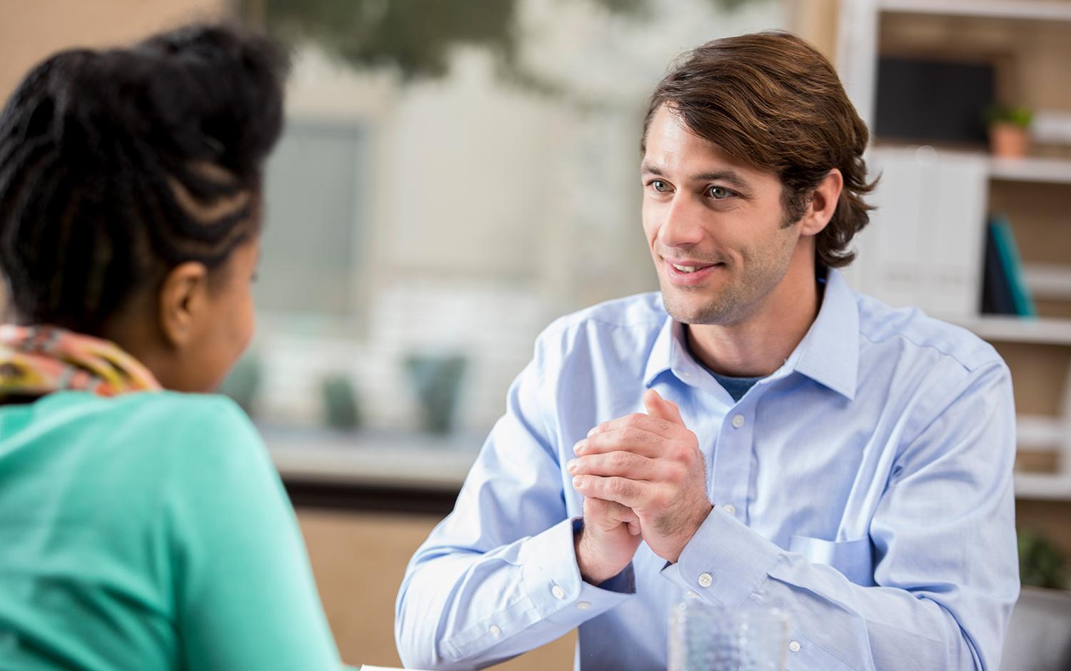 The image depicts two individuals sitting across from each other at a table. The person on the right appears to be gesturing with clasped hands, suggesting a conversation or discussion. The setting seems to be indoors, possibly an office or casual meeting space, with shelves and office supplies in the background.