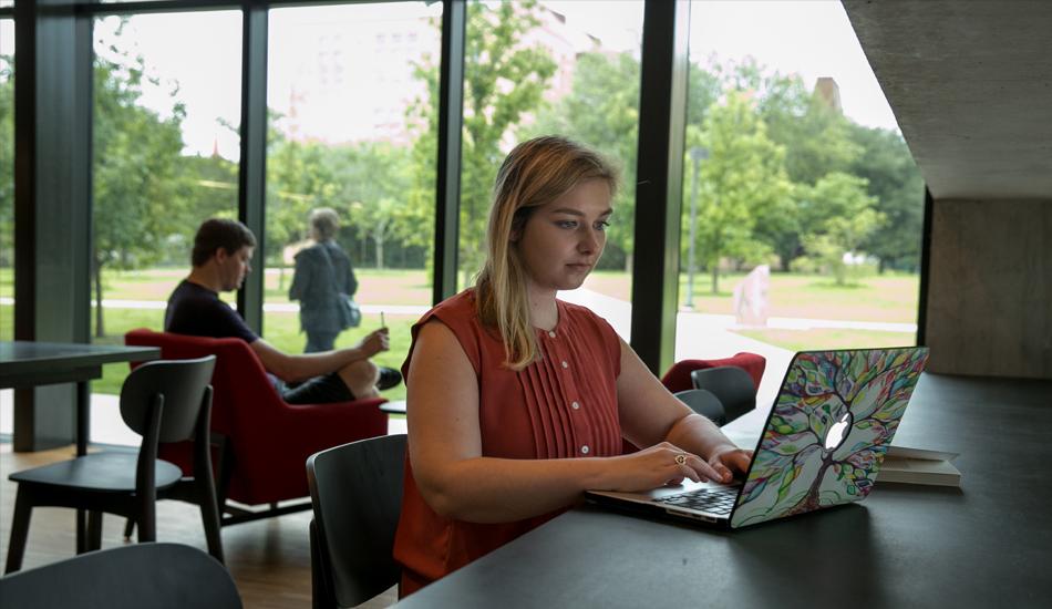 The image shows a young woman with blonde hair, wearing a sleeveless rust-colored blouse, working on a laptop in a modern, spacious room with large windows. Her laptop has a colorful tree design on the cover. She is seated at a long table, focused on her work. In the background, a man is seated in a red chair reading, while another person walks outside in a lush green park. The setting appears to be a quiet, well-lit study or lounge area with a view of the outdoors.