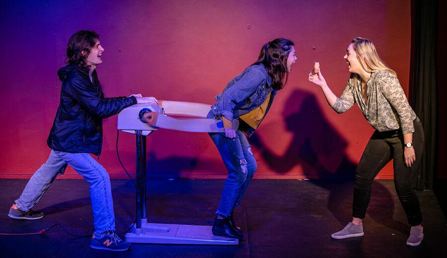 Three students participate in an improv scene with props while standing in front of a blue, red and purple background.