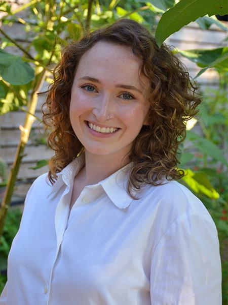 Kathryn Brouillette wears a white button up blouse. Greenery is in the background.