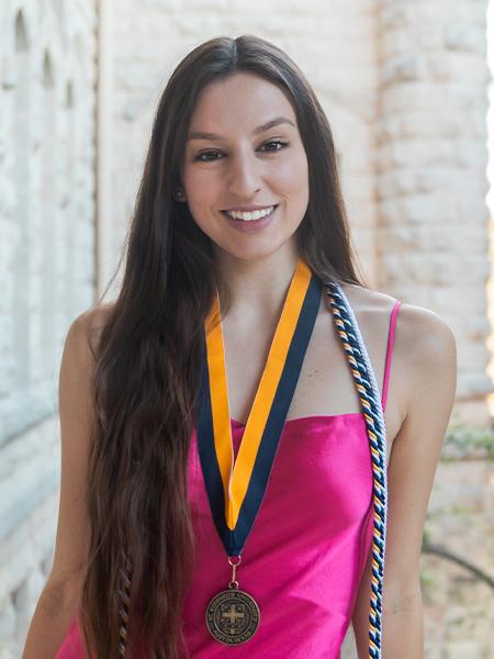 Marisa Davis wears a pink dress, a medallion and cords. Main Building is in the background.
