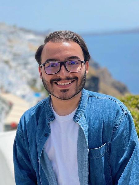 Nic Carrillo wears a white t shirt and a blue denim button up shirt and glasses. A coastal view is in the background.