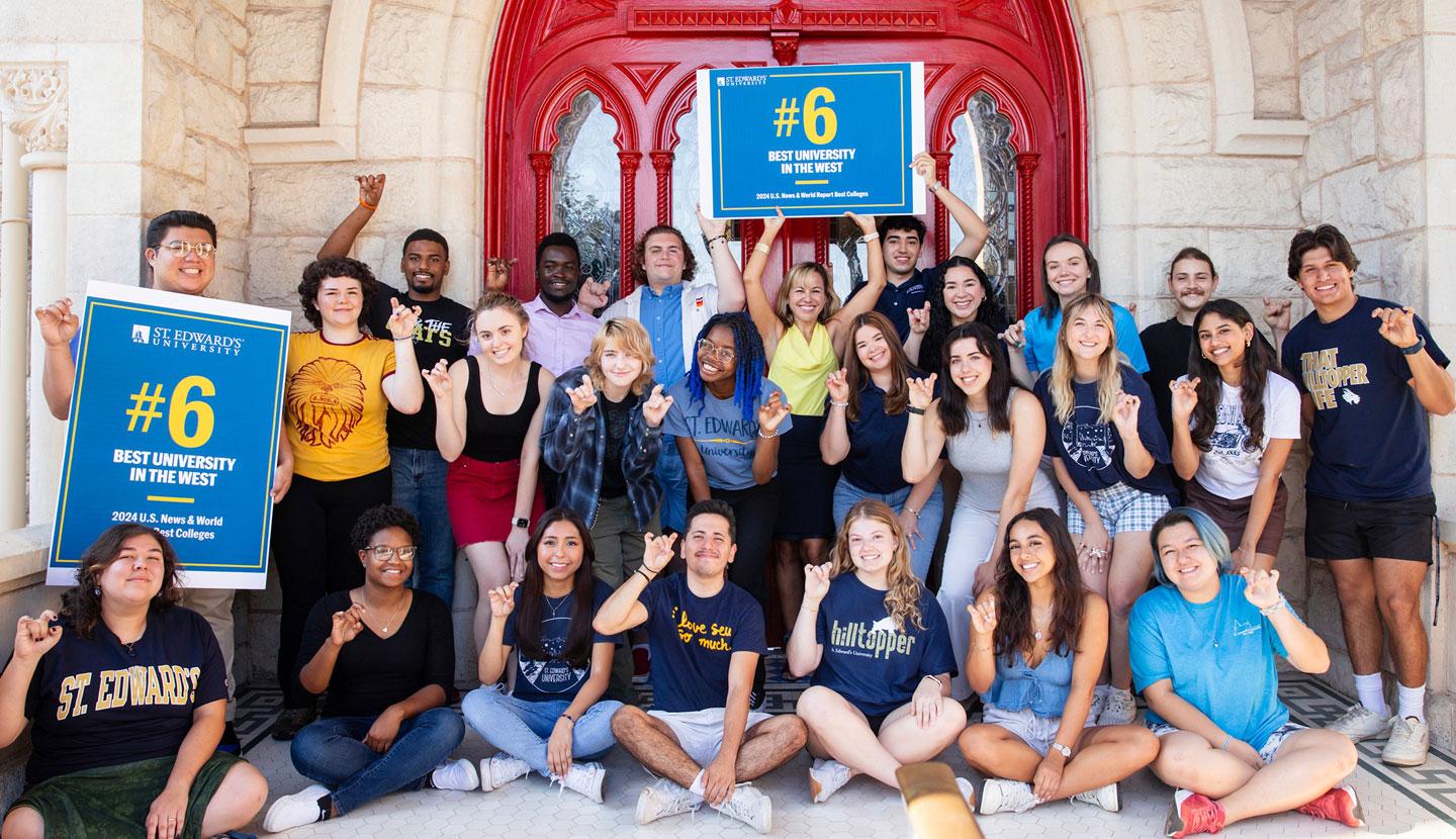 The image features a group of individuals posing in front of a building with a red door. Several people are holding blue signs with yellow and white text. One sign reads "#6 BEST UNIVERSITY IN THE WEST" and another "ST. EDWARD'S UNIVERSITY."