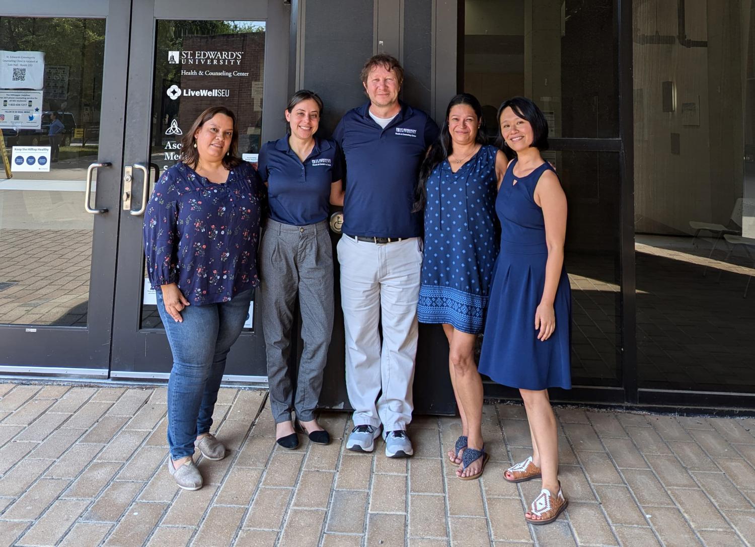 Five people standing in front of a glass door with signage for St. Edward's University Health & Counseling Center. The group consists of three women on the left and two women on the right, all wearing casual or business-casual attire in shades of blue and gray. They appear to be smiling and posing for a group photo. The background shows the entrance to the building with various logos and notices on the glass door.
